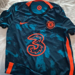 Chelsea FC Nike bortatröja blå/orange - Chelsea FC bortatröja från Nike med unikt blått och mörkblått mönster, orange detaljer och klubbmärke. Stort orange sponsortryck på bröstet och orange insida på kragen. Tillverkad i lätt och ventilerande material, perfekt för match eller träning.