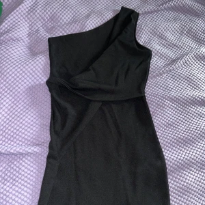 Svart one shoulder kortklänning - Snygg svart klänning med one shoulder-design och figurnära passform. Klänningen är kort (midi) och har en stilren look utan mönster eller extra detaljer. Perfekt för dig som gillar minimalistisk stil och vill sticka ut med en asymmetrisk siluett.
