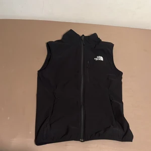 Svart väst från The North Face - Svart väst från The North Face med hel dragkedja framtill och två sidofickor. Västen har en hög krage och logga på både bröstet och ryggen. Perfekt för lager-på-lager och outdoor-vibe.