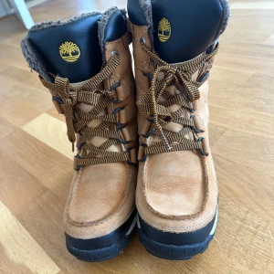 Timberland fodrade boots i skinn - Säljer ett par snygga Timberland boots i brunt och svart skinn med grov sula och snörning. Insidan är fodrad med mjukt, brunt material som håller fötterna varma. Perfekt för kalla dagar och outdoor vibes. Klassisk Timberland-logga på plösen och sidan.