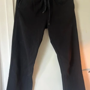Svarta mjukisbyxor från Gina Tricot - Svarta mjukisbyxor från Gina Tricot med elastisk midja och snörning framtill. Byxorna har en loose passform och är gjorda i mjukt polyester-material. Perfekta för chill dagar eller när du vill vara extra bekväm. Snyggt basic-plagg som funkar till allt.