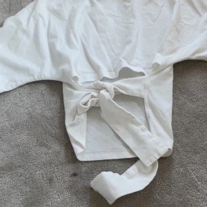 Vit croppad topp med knyt från Nelly - Säljer en vit croppad topp från Nelly i mjuk bomull. Toppen har rund halsringning och långa ärmar, samt en snygg knytning framtill som ger en cool och trendig look. Perfekt för dig som gillar stilrena och enkla plagg med en twist.