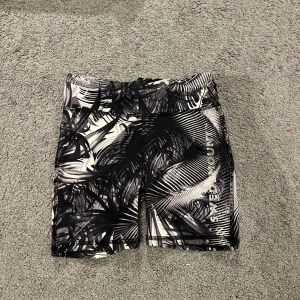 Svarta och vita träningsshorts Swedemount - Snygga träningsshorts från Swedemount i svart och vitt med grafiskt mönster av palmblad. De har en tight passform och är gjorda i stretchigt syntetmaterial som andas. Perfekta för sport eller gym och har en bekväm midja som sitter på plats under aktivitet.
