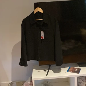 Svart overshirt från Zara, storlek S - Stilren svart overshirt från Zara i storlek S. Skjortan har klassisk krage, raka axlar och stora bröstfickor. Materialet är mjukt och lätt polyester, perfekt för lager-på-lager. Stängs med knappar framtill och har långa ärmar. Snygg och enkel att matcha.