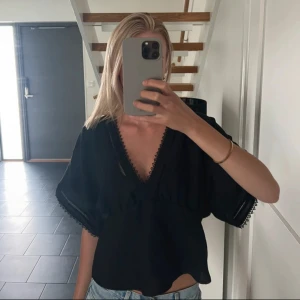 Svart v-ringad blus med spetsdetaljer - Svart blus med djup v-ringning och fina spetsdetaljer längs urringning och ärmslut. Modellen är loose fit med korta, vida ärmar och lätt croppad passform. 