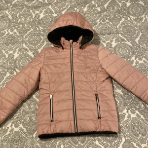 Ljusrosa quiltad pufferjacka med huva - Säljer en ljusrosa quiltad pufferjacka med huva och svarta detaljer. Jackan har två fickor med dragkedja, hel dragkedja framtill och svart foder på insidan. Perfekt för kyliga dagar och riktigt snygg till jeans eller mjukis.