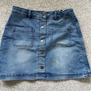 Blå jeanskjol från Tommy Hilfiger - Snygg blå jeanskjol från Tommy Hilfiger med raka linjer och två stora fickor framtill. Kjolen har knappar längs hela framsidan och bälteshällor i midjan. Perfekt för dig som gillar klassisk denimstil med coola detaljer.