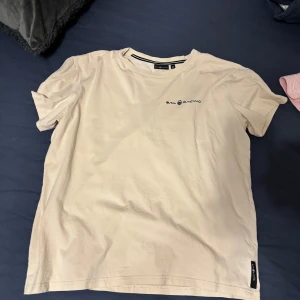 Beige t-shirt från Sail Racing - Snygg beige t-shirt från Sail Racing i mjuk bomull. Klassisk passform med rund hals och diskret logga på bröstet samt liten etikett i nederkant. Perfekt basic-plagg med stilren look och neutrala färger.