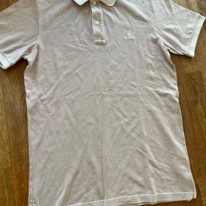 Beige pikétröja från GANT - Klassisk beige pikétröja från GANT i storlek S. Tröjan har korta ärmar, krage och knäppning med två knappar framtill. Broderad GANT-logga på bröstet. Tillverkad i mjuk bomull som känns skön mot huden. Perfekt för en clean och stilren look.