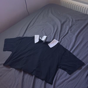 Svart croppad topp med vit krage Kappahl - Snygg svart croppad topp från Kappahl med kontrasterande vit krage. Oversized passform och korta ärmar ger en trendig look. Perfekt för dig som gillar stilrena detaljer och vill sticka ut med en klassisk touch.