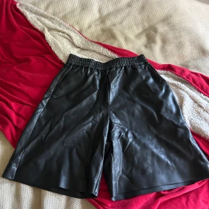 Svarta skinnshorts från Lindex - Snygga svarta shorts från Lindex i skinnimitation med elastisk midja. Modellen är loose fit och har en stilren, rak form som ger en cool vibe. Perfekta att matcha med en oversized t-shirt eller hoodie för en trendig look.