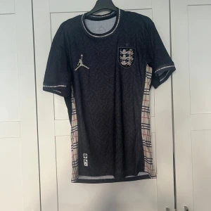 Svart England x Jordan fotbollströja - Snygg svart England fotbollströja från Jordan i storlek M men den är liten i storleken så den passar S med och köpt från fotbollsmarknaden .