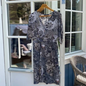 Mönstrad klänning från Ilse Jacobsen M - Snygg klänning från Ilse Jacobsen med svart och grått paisleymönster. Modellen har v-ringning, trekvartsärm och knälång passform. Perfekt för dig som gillar unika prints och en bekväm men stilren look. Snyggt fall och bekvämt tyg!