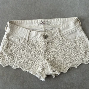 Vita spetsdetaljerade shorts från Hollister - Superfina vita shorts från Hollister med spets framtill och råa kanter. Klassisk femficksmodell med knapp och dragkedja. Lågmidja och jättefina! Perfekta för sommaren och ger en romantisk touch till outfiten.