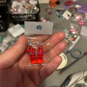 Örhängen med läskflaskor - Unika örhängen formade som små röda läskflaskor. Perfekta för dig som vill sticka ut och addera en rolig detalj till din stil. Säljs i par och har silvriga krokar.