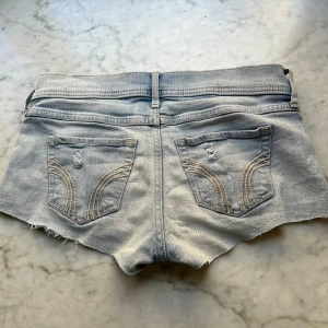Lågmidjade shorts - Ljusblå jeansshorts från hollister med klassiska fickor bak och fram, råa och fransiga benslut samt knapp och dragkedja framtill. Snygga detaljer med slitningar och broderade sömmar på bakfickorna. Perfekta för varma dagar.