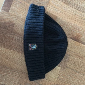 Svart ribbad mössa från Parajumpers - Säljer en svart ribbstickad mössa från Parajumpers med uppvikt kant och broderad logotyp framtill. Klassisk och stilren design som passar perfekt till kyliga dagar.