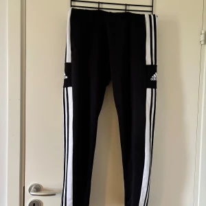 Svarta träningsbyxor från Adidas med vita ränder - Säljer ett par svarta träningsbyxor från Adidas med klassiska vita ränder längs sidorna och Adidas-logga på låret. Byxorna har en sportig look och är perfekta för träning eller chill. Snygg och enkel design.