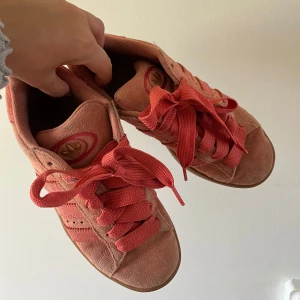 Rosa Adidas campus skor - Säljer ett par rosa Adidas campus sneakers i mocka med klassiska tre ränder på sidan och breda rosa skosnören. Skorna har rund tå och platt sula, perfekt för dig som gillar färg och vill sticka ut lite extra.