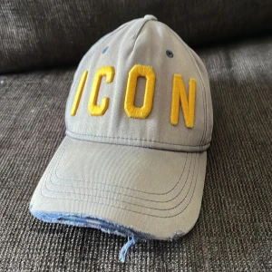 Grå/brun  keps från Dsquared2 med gul brodyr - Grå keps med böjd skärm från Dsquared2. Framtill står det 'ICON' i stora gula broderade bokstäver och baktill 'DSQUARED2'. Kepsen har justerbar rem bak 