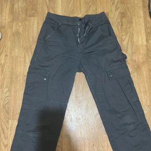 Mörkgrå cargopants med dragkedja - Säljer ett par mörkgrå cargopants med flera fickor på benen och klassisk dragkedja framtill. Byxorna har normal passform och är perfekta för en avslappnad stil. Snygga detaljer med knappar på fickorna och bälteshällor i midjan.