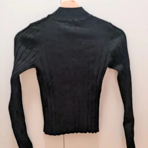 Svart ribbad polotröja - Säljer en svart ribbad polotröja med lång ärm och tight passform. Tröjan är croppad och har en hög krage som ger en stilren look. Perfekt att styla med jeans eller kjol för en trendig outfit.