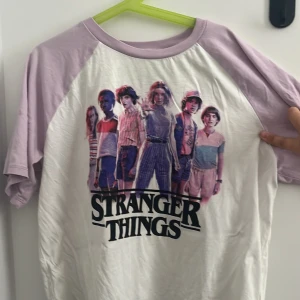 Stranger Things t-shirt från H&M - T-shirt med motiv från Stranger Things. Ljuslila ärmar och krage, vit framsida med tryck av karaktärerna och loggan. Perfekt för fans av serien och snygg till vardags.