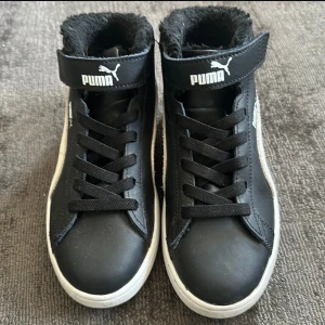 Svarta sneakers från Puma med fuskpälsfoder - Säljer ett par svarta Puma sneakers med vit sula och vit sidodetalj. Skorna har snörning, kardborreband upptill och är fodrade med mjuk fuskpäls. Innersulan är märkt med SoftFoam+ för extra komfort.