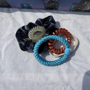 Flerpack hårsnoddar i olika material och färger - Säljer ett set med fyra hårsnoddar: en blå flätad med pärldetalj, en svart sammetsliknande scrunchie, en beige spiral, och en brun spiral. Perfekta för att variera din frisyr och stil.