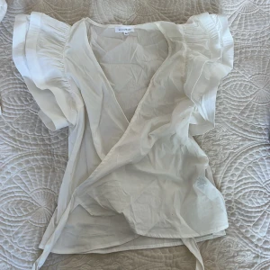 Vit omlottblus med volangärm från Lulumari - Säljer en vit omlottblus från Lulumari med korta volangärmar och knyt i midjan. Blusen har en luftig och feminin design, perfekt för varma dagar. Tunn och mjuk bomullskänsla.