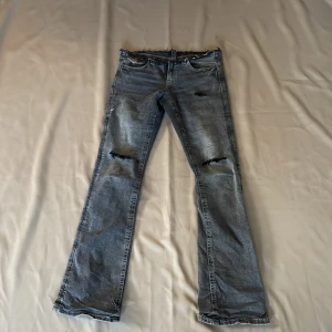 Grå slitna bootcut jeans - Säljer ett par grå bootcut jeans med slitningar på knäna och rå kant i midjan. Jeansen har fem fickor och dekorativa sömmar på bakfickorna. Perfekta för en avslappnad och trendig look. I storlek s/xs 