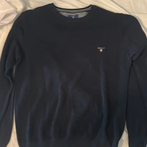 Mörkblå stickad tröja från GANT - Säljer en klassisk mörkblå stickad tröja från GANT med rund halsringning och diskret logga broderad på bröstet. Perfekt för en stilren och enkel look. Tröjan har långa ärmar och ribbade muddar.