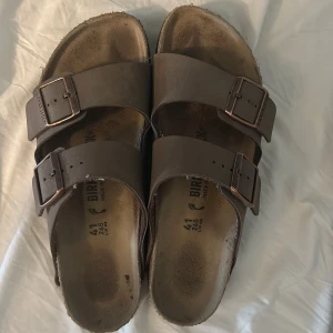 Bruna tofflor  från Birkenstock - Klassiska bruna sandaler från Birkenstock med två breda remmar och justerbara metallspännen. Fotbädden är anatomiskt utformad och sulan har ett vågigt mönster för bra grepp. Perfekta för varma dagar och enkel att matcha med olika outfits.