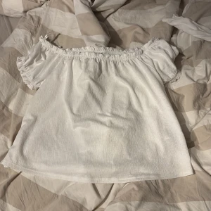 Vit offshoulder topp från Gina Tricot - Säljer en söt vit offshoulder topp från Gina Tricot i storlek M. Toppen har resår vid axlarna och korta ärmar med volangkant. Perfekt för varma dagar och enkel att matcha med olika outfits. Har inte använt denna. 