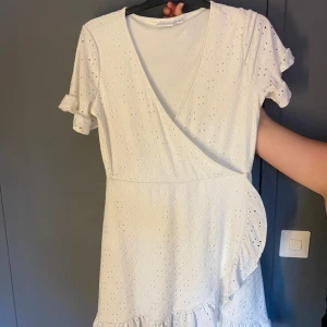 Studentklänning vit klänning  - Söt vit kortklänning från VILA med omlott-design, volangdetaljer och korta ärmar med rysch. Klänningen har broderat hålmönster och v-ringning. Perfekt för studenten eller midsommar.