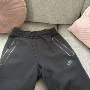 Svarta mjukisbyxor från Nike med dragkedjor - Svarta mjukisbyxor från Nike med justerbar midja och dragkedjor på fickorna. Byxorna har snörning i midjan och dragsko vid benslut för extra passform. Perfekta för en sportig och avslappnad stil.