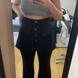 Svarta byxor med kjoldetalj och knappar - Säljer ett par svarta byxor med unik kjoldetalj framtill och stora dekorativa knappar. Byxorna har fickor framtill och är utsvängda nertill. Perfekta för dig som vill sticka ut med din stil.