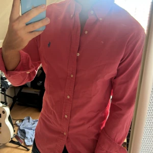 Rosa skjorta från Ralph Lauren - Säljer en klassisk rosa skjorta från Ralph Lauren med blått broderat logotyp på bröstet. Skjortan har lång ärm, knappar framtill och krage. Perfekt för dig som gillar stilrena plagg med en färgklick.