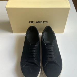 Svarta sneakers från Axel Arigato - Snygga svarta sneakers från Axel Arigato med klassisk design och svarta snören. Skorna har en platt vit sula och rund tå, perfekt för en stilren vardagslook. Levereras med originalkartong.