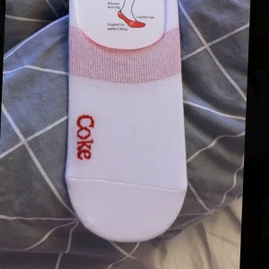 Vita låga strumpor med röd Coca-Cola text - Säljer ett par vita låga strumpor med röd Coca-Cola text på sidan. Strumporna har silikon på insidan för att sitta kvar på foten och är designade för att inte synas i skorna. Perfekta till sneakers eller låga skor.