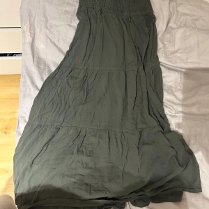 Grön långkjol med smock i midjan - Säljer en grön långklänning med smock upptill och volangdetaljer. Klänningen har en avslappnad passform och är perfekt för varma dagar. Passar lika bra till stranden som till sommarens alla tillfällen.
