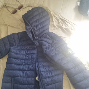 Blå pufferjacka från Stone Island - Säljer en blå pufferjacka från Stone Island med huva och dragkedja. Jackan har den klassiska loggan på ärmen och quiltad design. Perfekt för kyligare dagar och riktigt snygg till streetwear-stilen. Kom privat ifall ni vill veta mer info fråga mig! Helt ny jacka förutom stone island märket some lite slitet efter den tvättats med andra kläder utan sin jacka ens men jackan är helt ny, använd 1 gång!