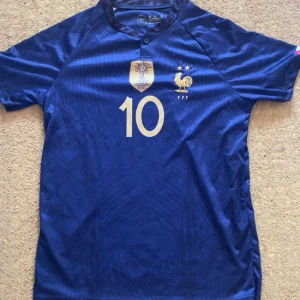 Frankrike fotbollströja Mbappé #10 - Säljer en blå fransk landslagströja med guldiga detaljer, nummer 10 och Mbappé på ryggen. Tröjan har korta ärmar, två stjärnor ovanför tuppen och FIFA-märke på bröstet. Perfekt för dig som älskar fotboll och vill stötta Frankrike!