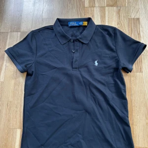 Svart pikétröja från Polo Ralph Lauren - Klassisk svart pikétröja från Polo Ralph Lauren med broderad logga på bröstet. Tröjan har krage, tre knappar framtill och är kortärmad. Perfekt för en stilren och tidlös look. Sitter litet passar M men S och mindre bättre