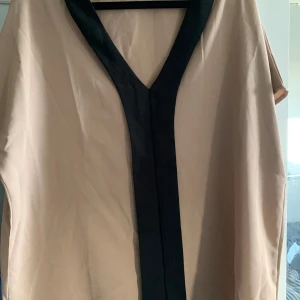 Beige blus med svart kant - Säljer en beige blus med svart v-ringad kant och lös passform. Blusen  har korta ärmar och är tillverkad i ett slätt material, perfekt för en stilren look.