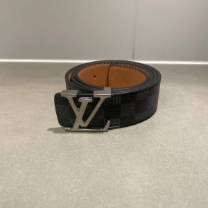Svart LV bälte - Säljer ett svart bälte från Louis Vuitton med klassiskt rutigt mönster och silverfärgat LV-spänne. Bältet har en stilren design och insida i ljusbrunt skinn. Perfekt accessoar för att lyfta din outfit.