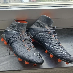 Nike hypervenom  - Säljer ett par mörkgråa Nike fotbollsskor med hög stickad strumpa och orangea dobbar. Skorna har snörning och en texturerad ovansida för bättre bollkontroll. Snygga detaljer i vitt och orange. Perfekta för dig som vill sticka ut på planen. Skick 9/10