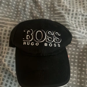 Svart keps från Hugo Boss - Snygg svart keps från Hugo Boss med broderad logga framtill och justerbart spänne baktill. Kepsen är tillverkad i 80% polyester och 20% bomull och har klassisk böjd skärm. Perfekt accessoar för en stilren look.