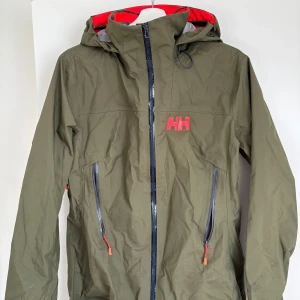 Grön vindjacka från Helly Hansen - Snygg grön vindjacka från Helly Hansen med huva och röda detaljer. Jackan har dragkedja framtill, två snedställda fickor med dragkedja och justerbar huva. Perfekt för utomhusaktiviteter när det blåser.