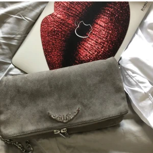 Grå axelväska med kedja från Zadig Voltaire  - Säljer en snygg grå axelväska i mocka med silverfärgad kedja från Zadig Voltaire . Väskan har dragkedja och passar perfekt när du vill ha med det viktigaste. Stilren och enkel att matcha till olika outfits.